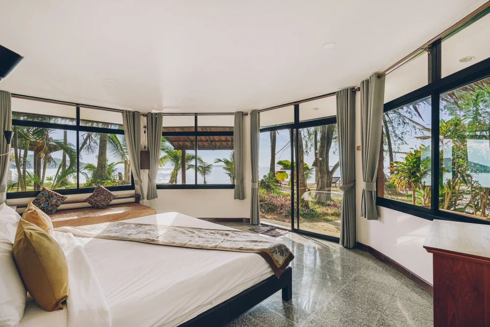 Bright bungalow bedroom with floor-to-ceiling windows opening to greenery and a sea glimpse.
