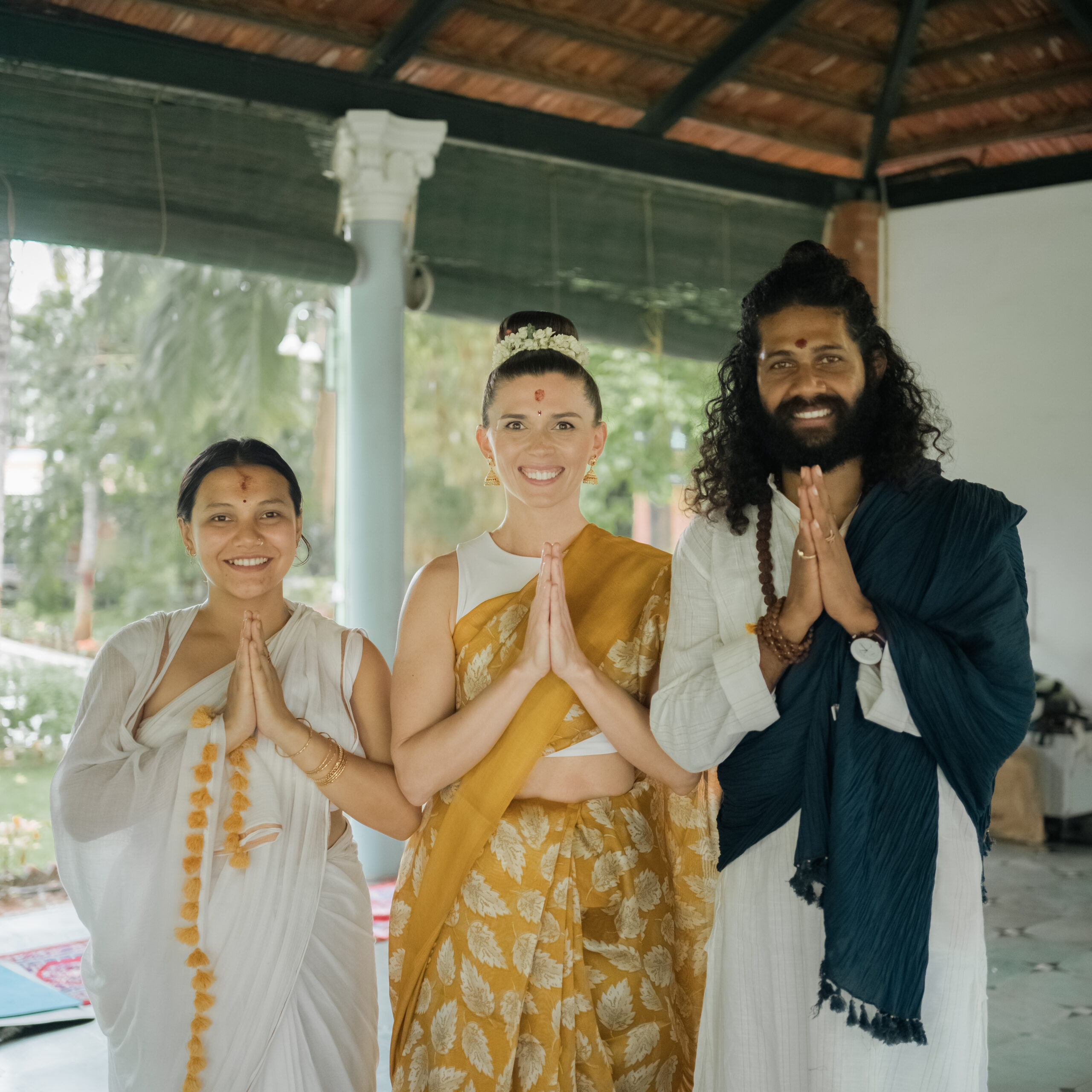 Three people in traditional Indian dress with marigold garlands greeting with namaste