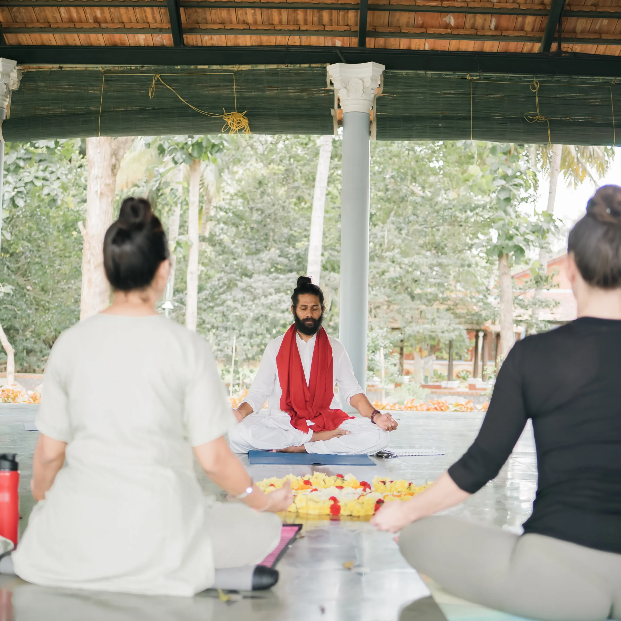 Students offering flowers to a teacher during a ceremony in an open-air yoga shala