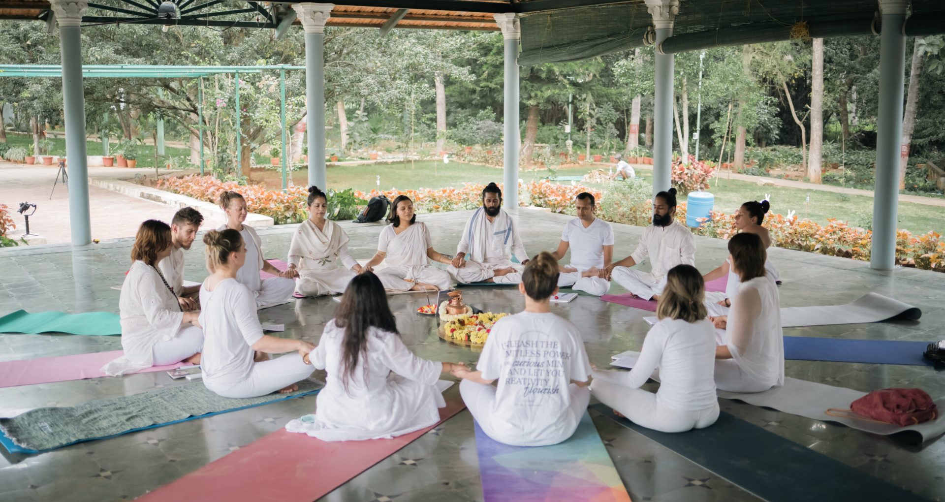 Yoga students seated on coloured mats in an open-air shala surrounded by greenery